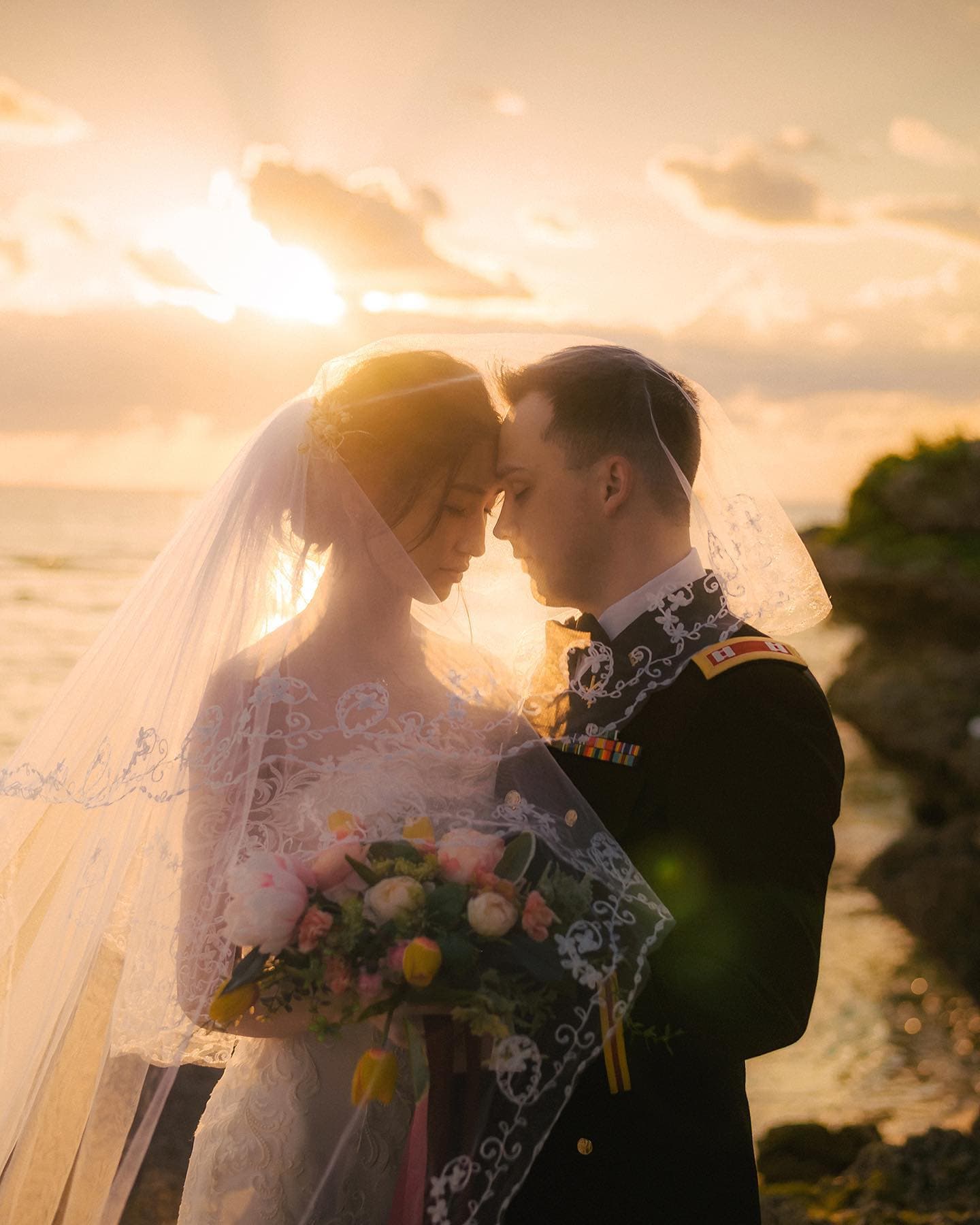 夕焼けの海辺でレースのベールに包まれて額を寄せる花嫁と礼装の新郎、沖縄