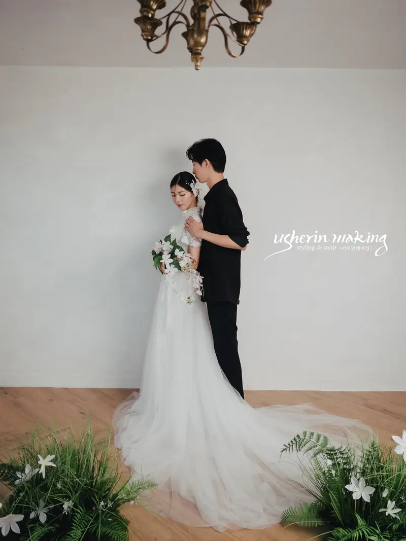 Groom embracing the bride from behind beneath a chandelier, white lilies at their feet