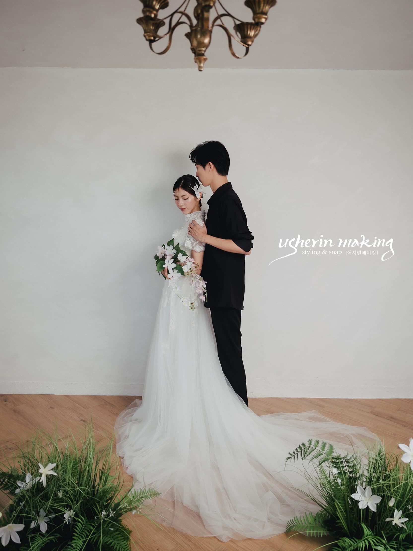 Groom embracing the bride from behind beneath a chandelier, white lilies at their feet