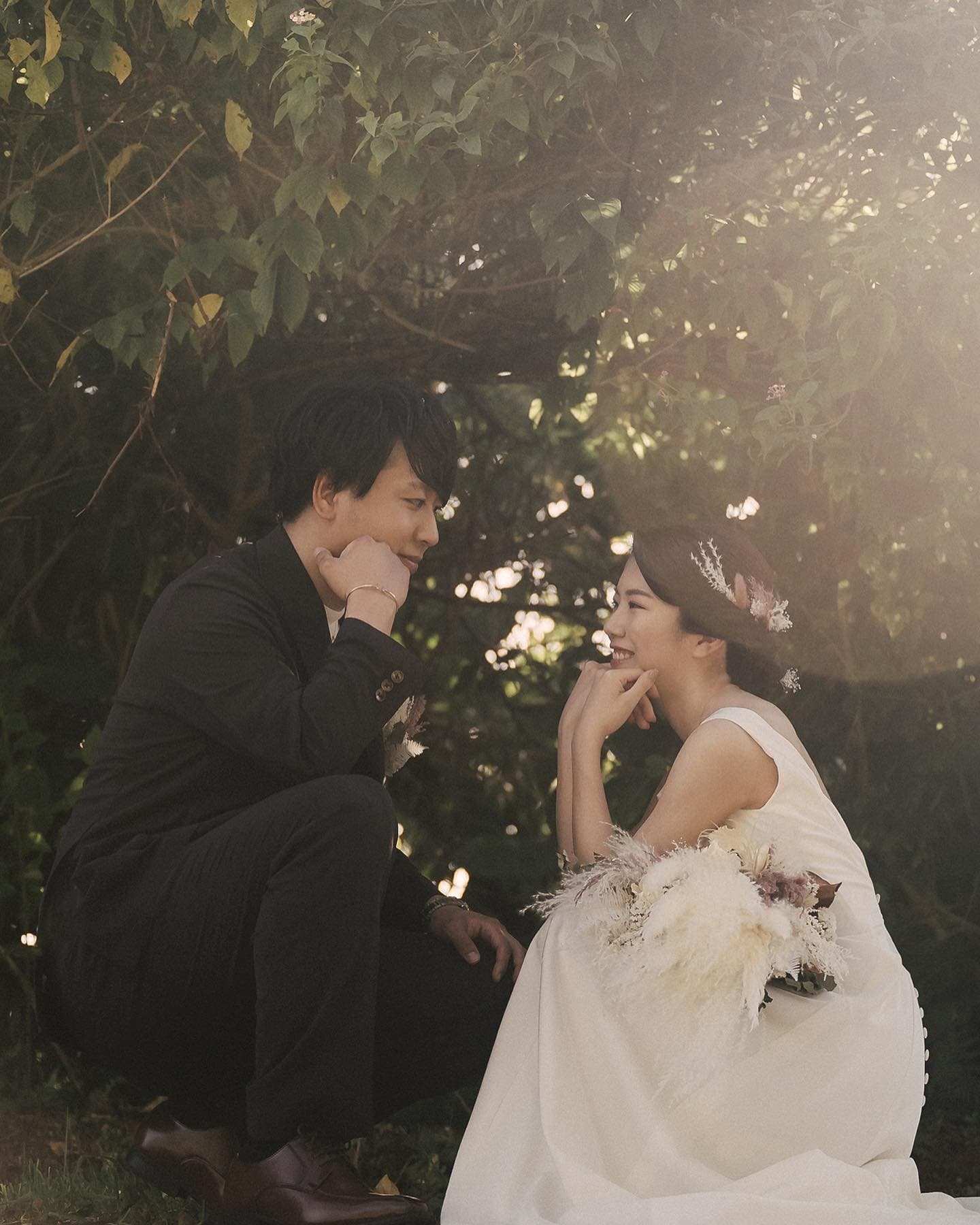 A bride and groom crouching face to face beneath a shady tree in Okinawa