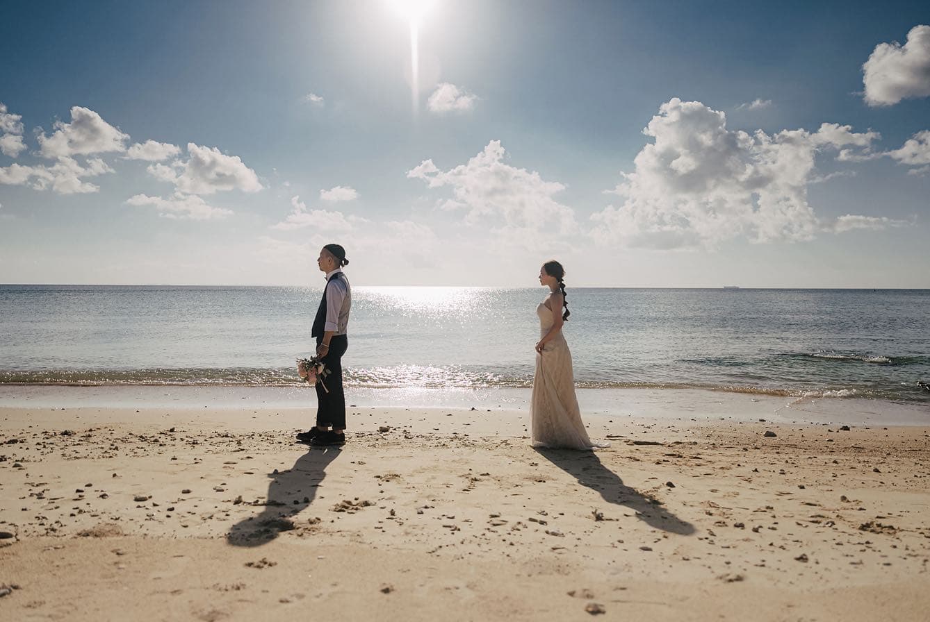 青空の下、沖縄のビーチで背中合わせに立つ新郎新婦