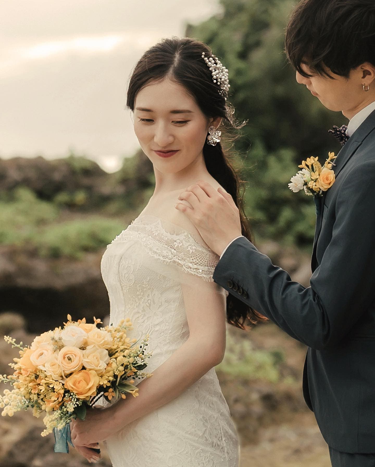 レースのドレスの花嫁がオレンジのバラのブーケを持ち、新郎が肩に手を添える