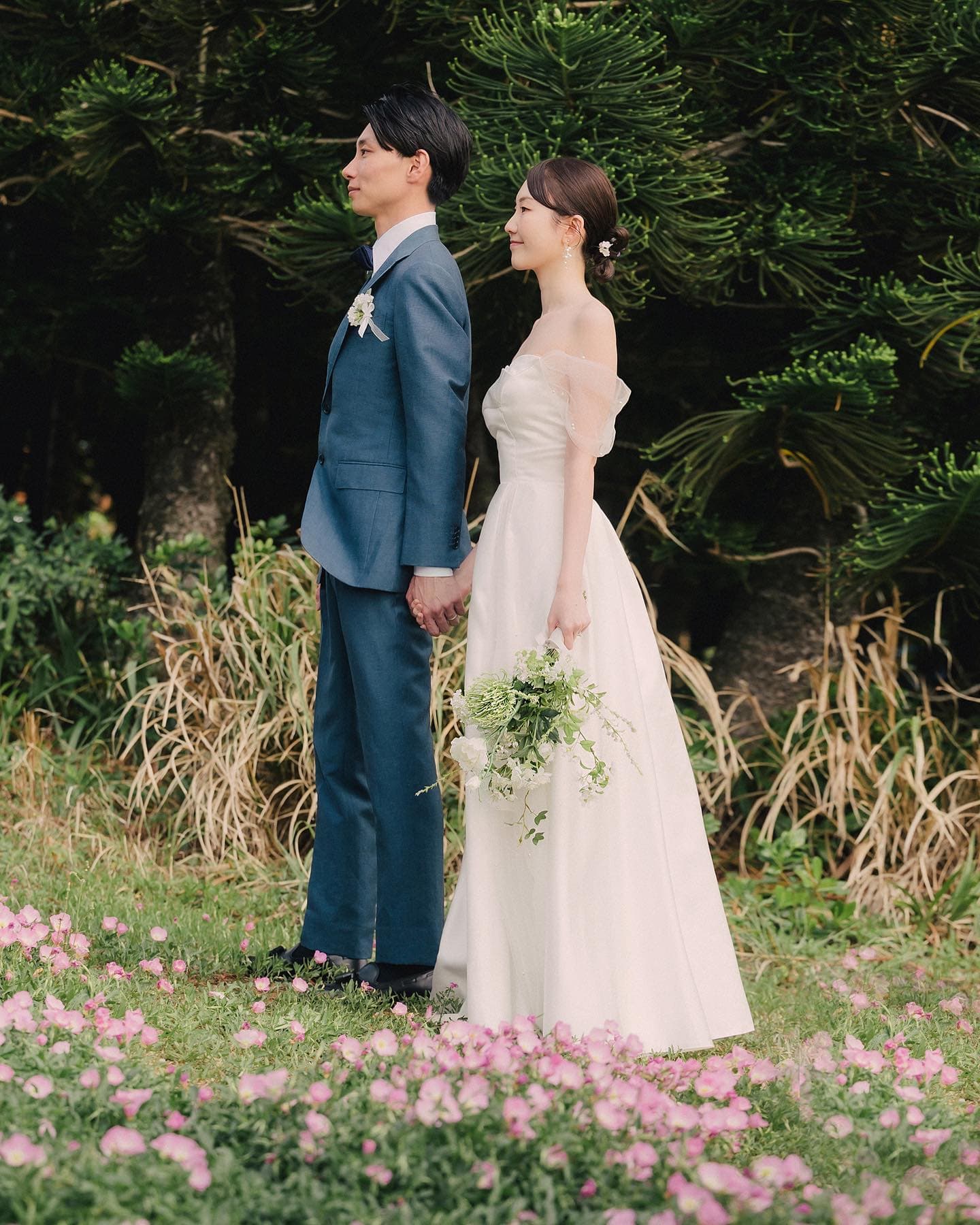 ピンクの花畑で手をつなぐネイビースーツの新郎とドレスの花嫁、沖縄