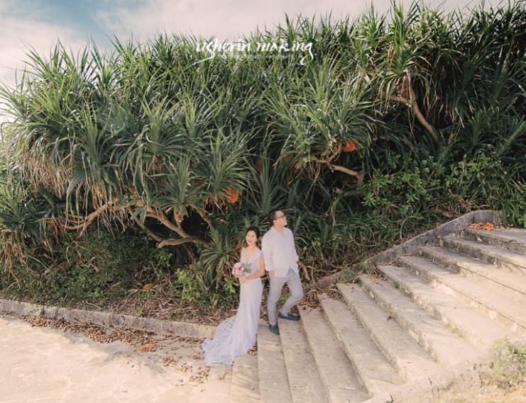 Couple in wedding attire on beachside stone steps under a blue Okinawa sky
