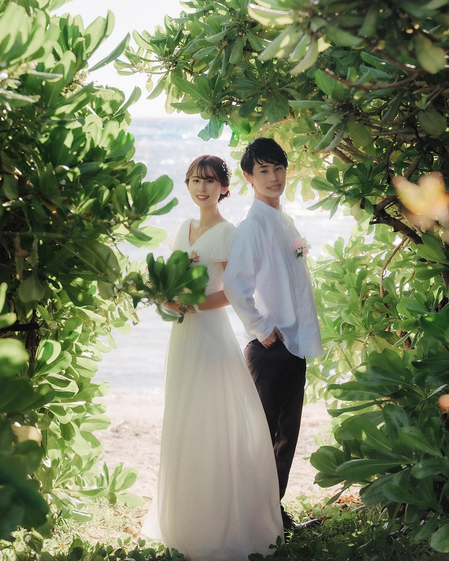 緑の木々の間で背中を合わせて立つ新郎新婦、沖縄の浜辺