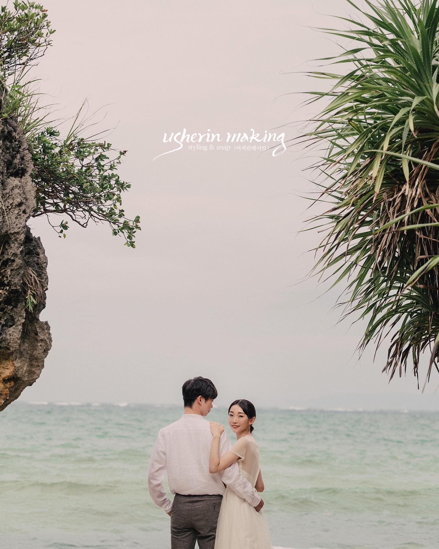 Couple in simple white outfits by a calm sea under an overcast Okinawa sky