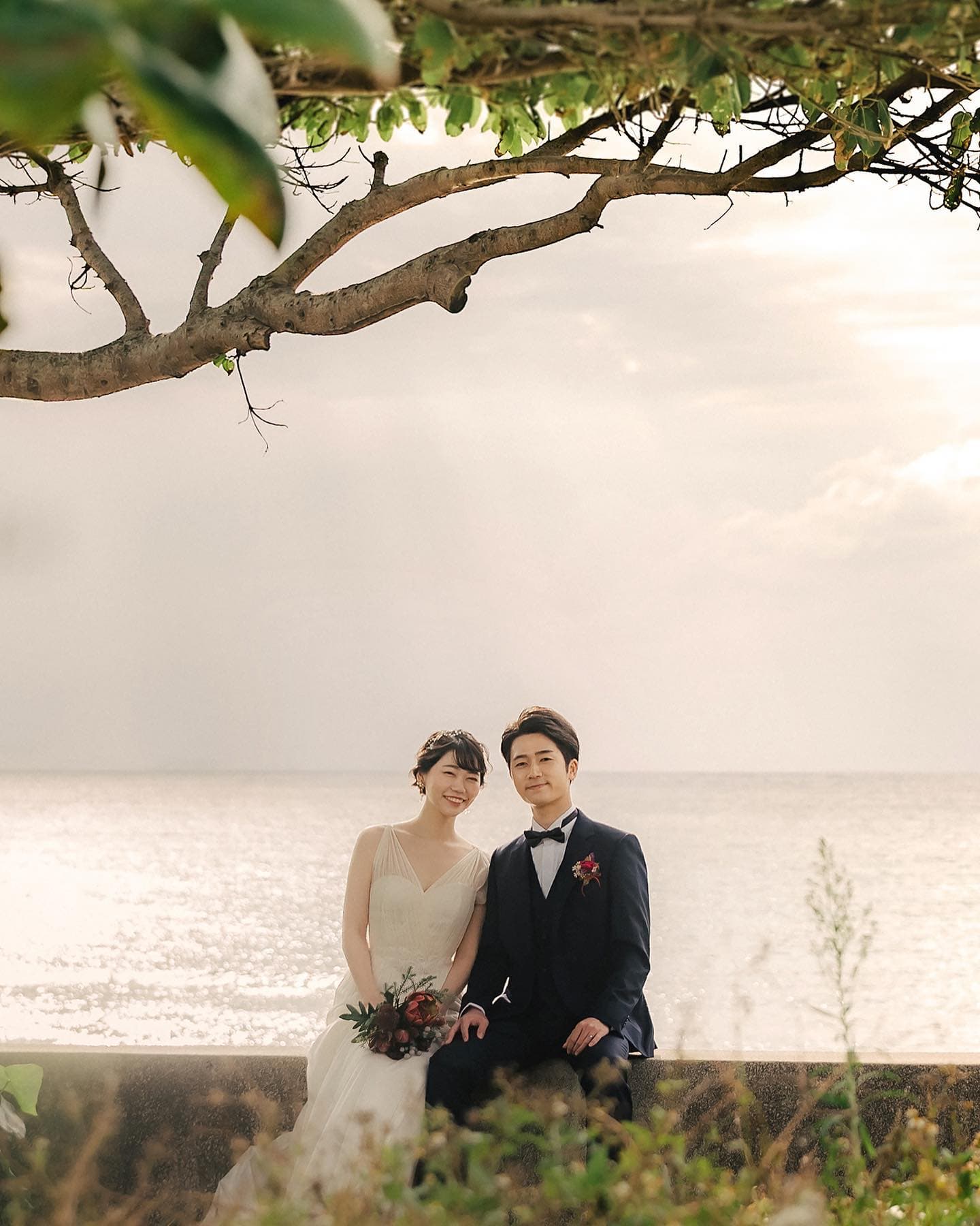 Bride and groom seated by a shimmering sea beneath an overhanging branch in Okinawa