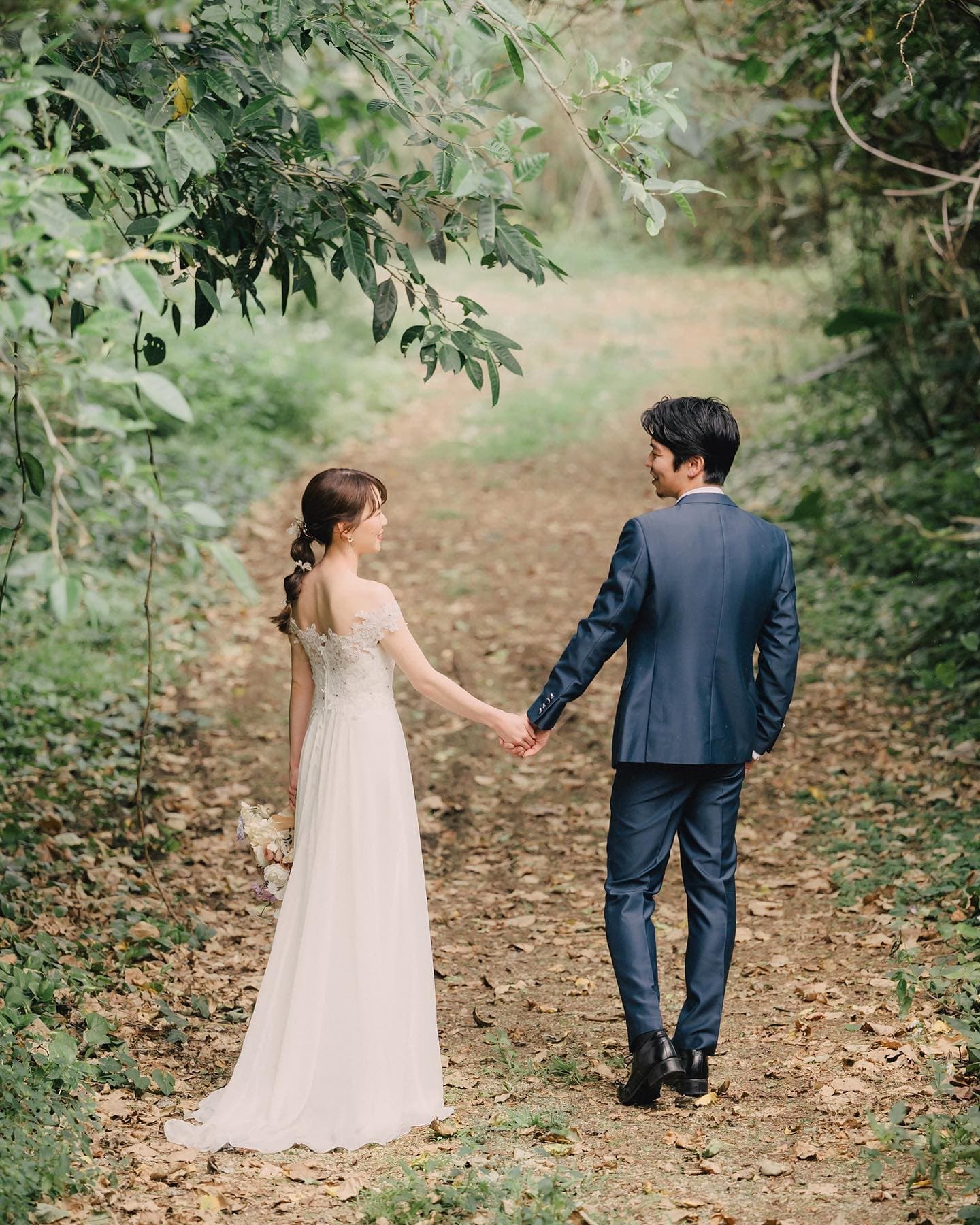 Couple in wedding attire strolling hand in hand down an Okinawan forest path