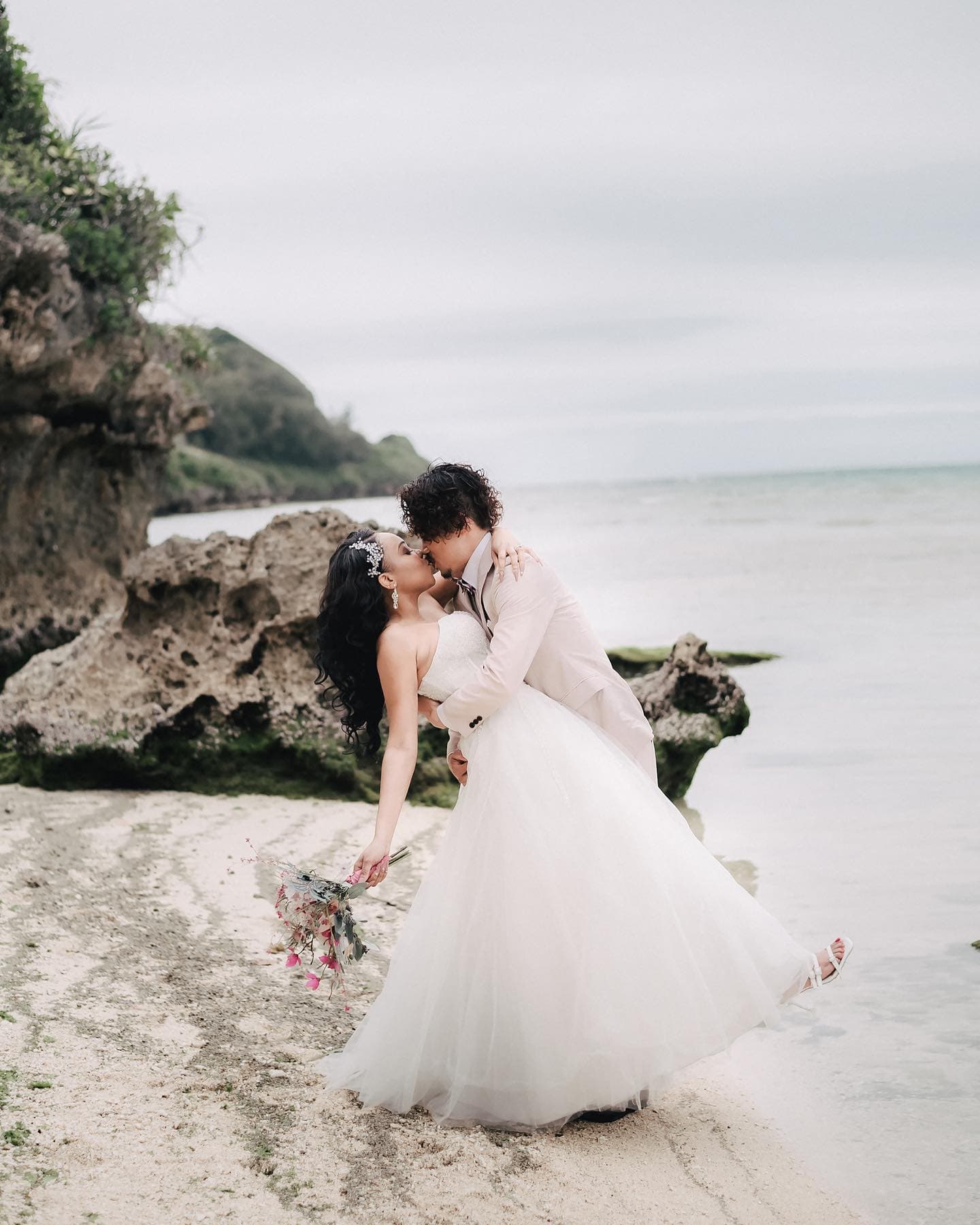 A couple kissing on an Okinawa beach by weathered rocks under a cloudy sky