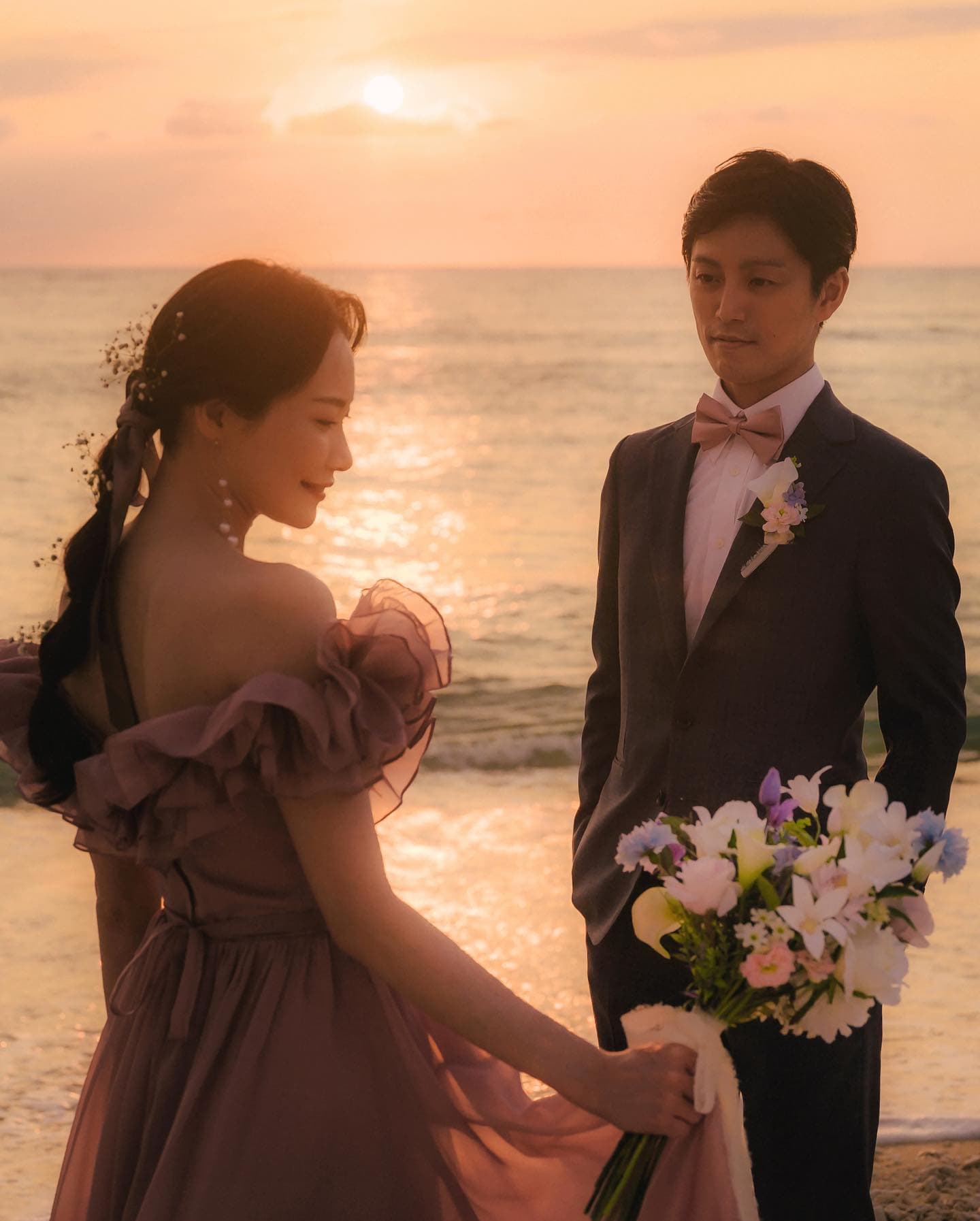 夕暮れの沖縄の海辺で、ピンクのドレスの花嫁を見つめる新郎