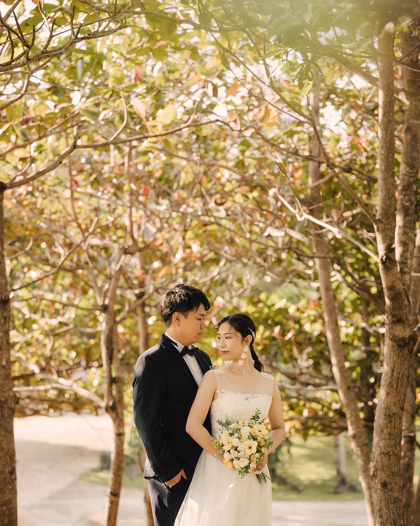 沖縄の木立の下で寄り添う新郎と黄色いブーケの新婦