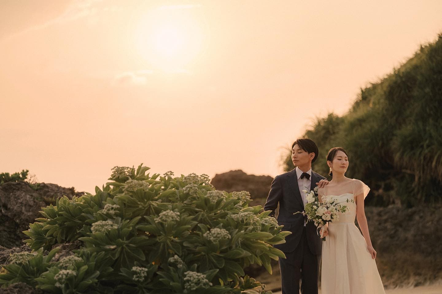 夕日に照らされた沖縄の海辺、白い花のそばに立つ新郎新婦