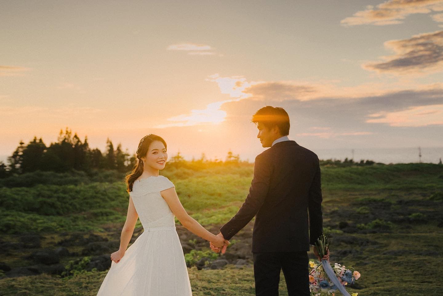 Bride glancing back with a smile as the couple walks through a sunset field