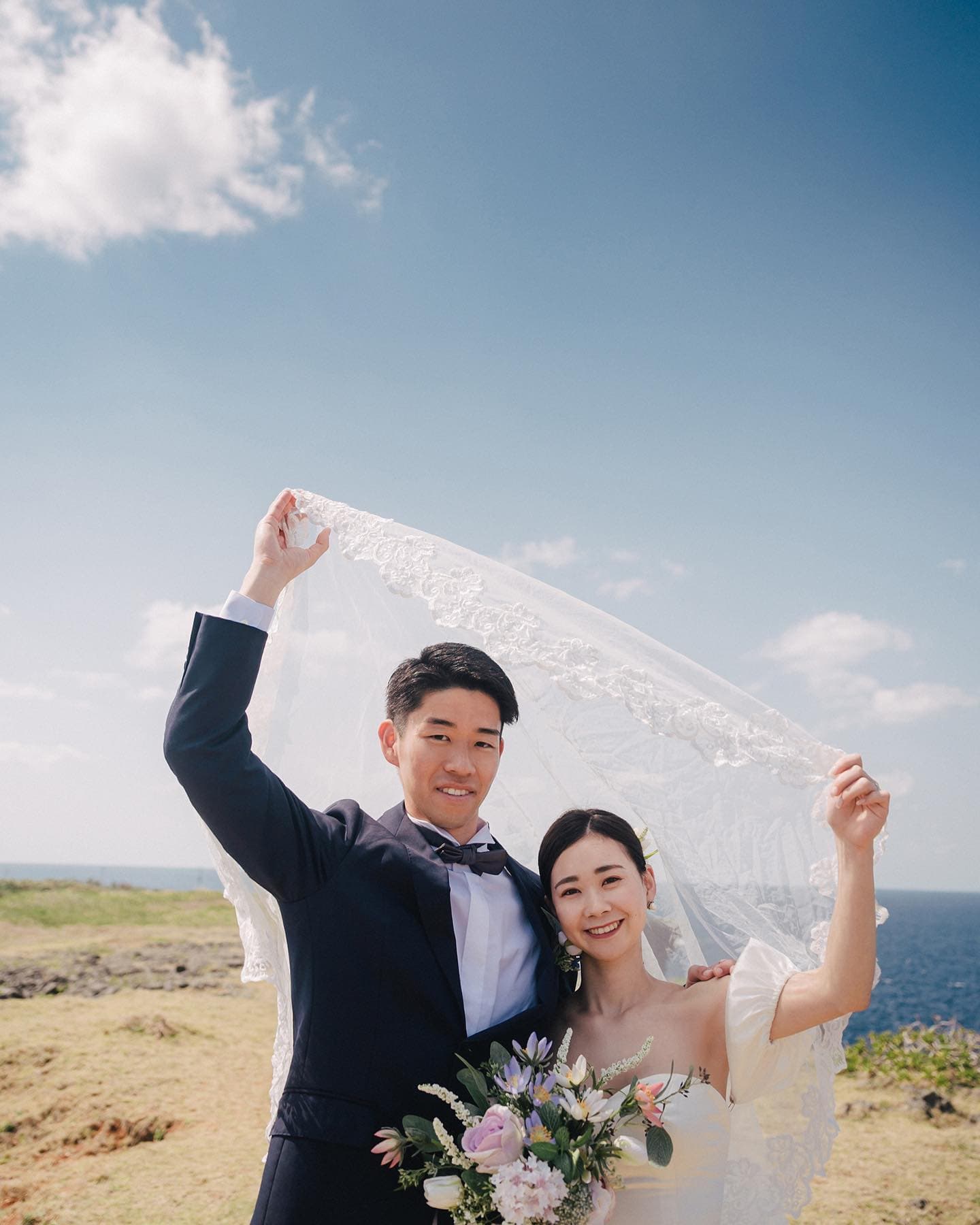 青空の下ベールを掲げて笑う新郎新婦、沖縄の岬