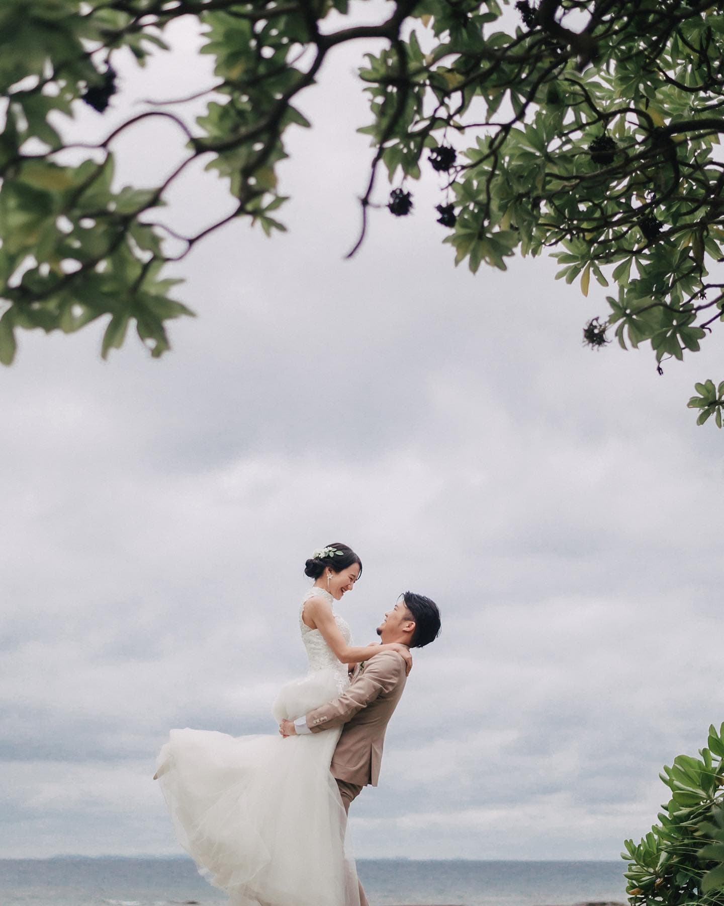 A groom lifts his bride by the sea under an overcast Okinawa sky