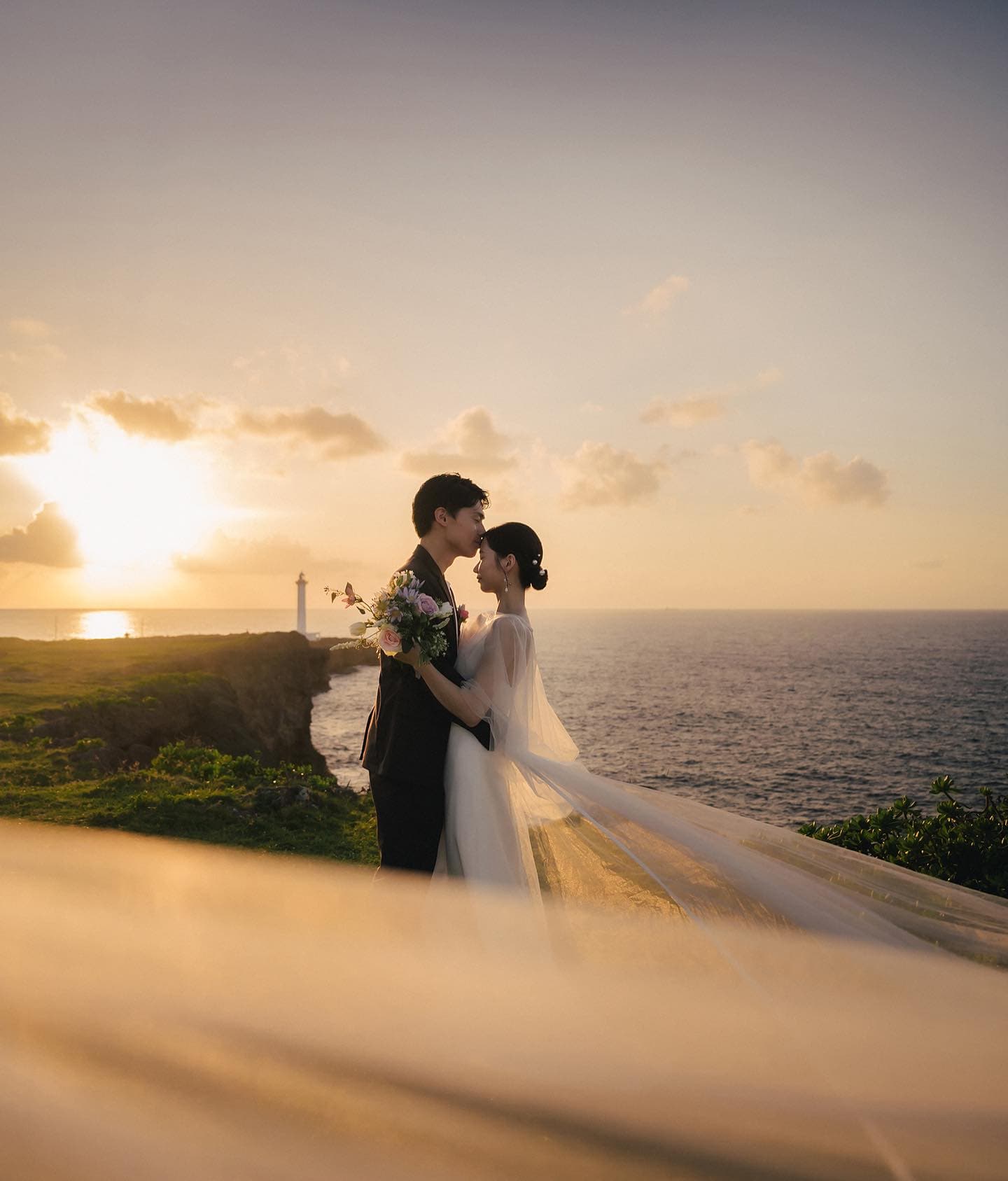 夕焼けの岬でヴェールが流れる中、新婦の額にキスする新郎、沖縄の灯台