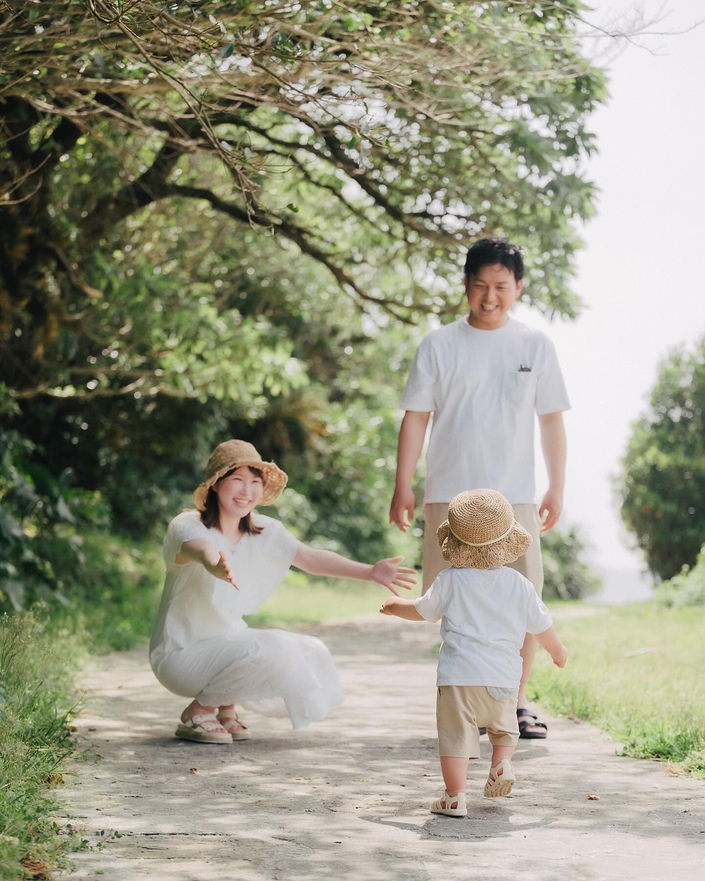 母の腕へ歩み寄る帽子の男の子と見守る父、緑の小道、沖縄の家族写真