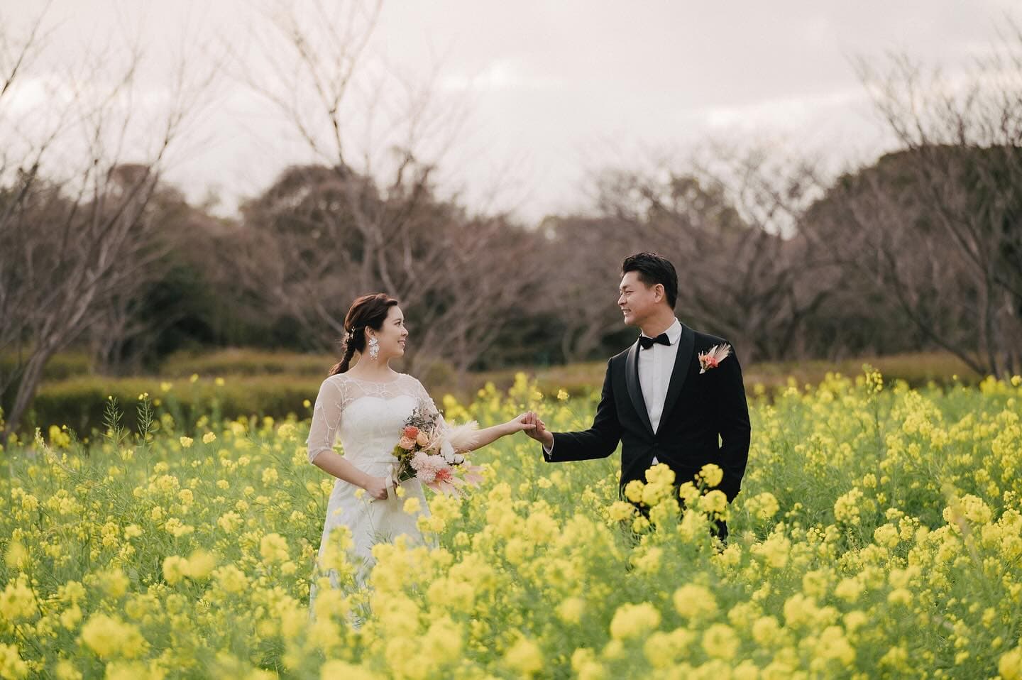 Couple holding hands in a yellow canola flower field under an overcast sky in Fukuoka