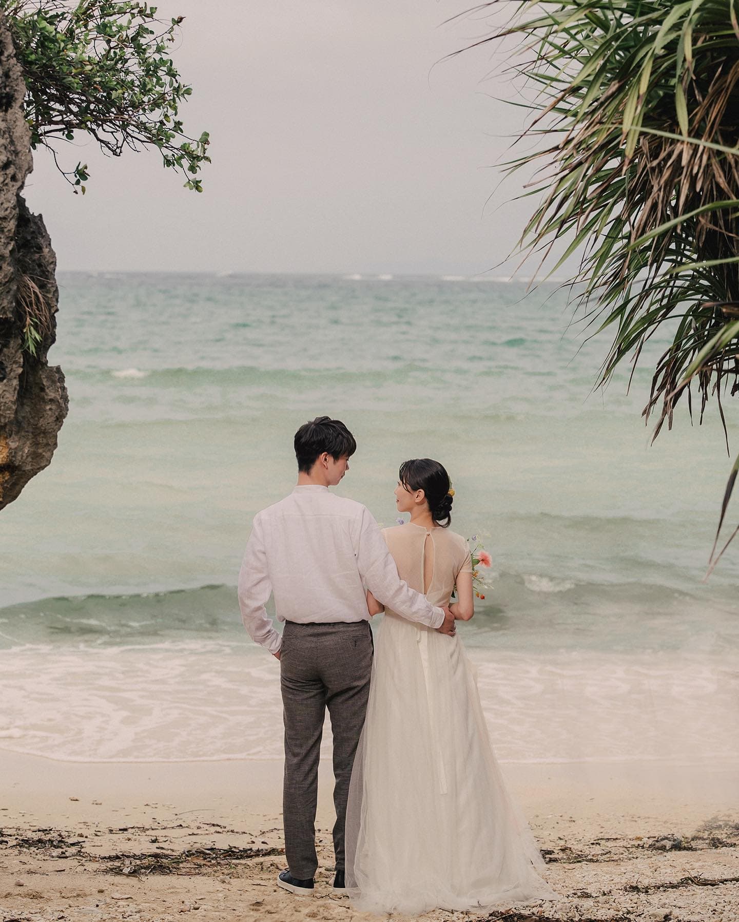 Couple standing arm in arm at the water's edge under a soft overcast sky in Okinawa