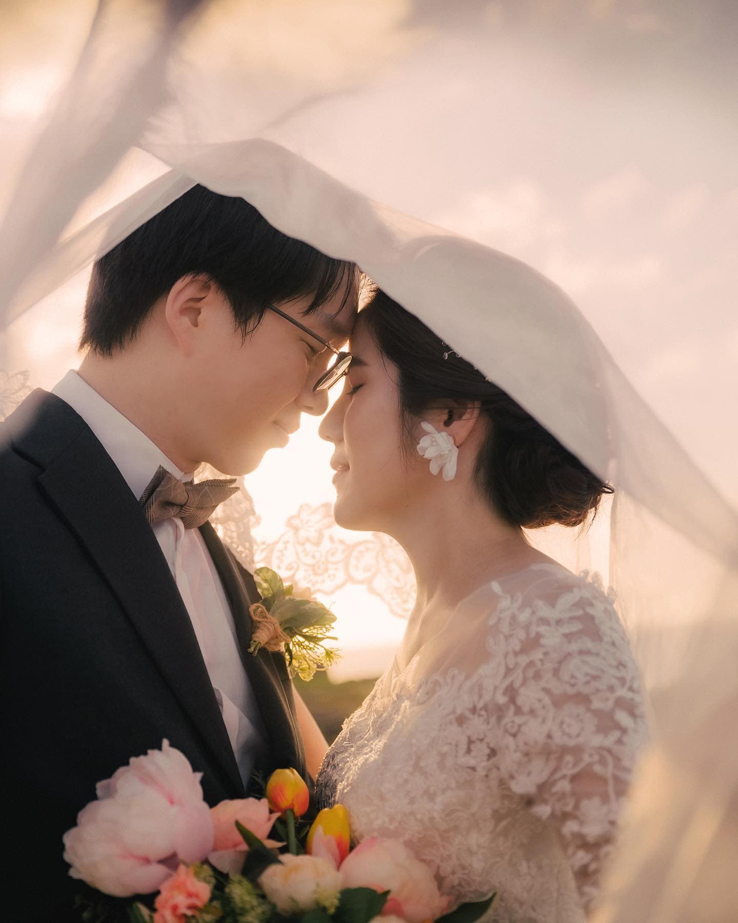 베일 아래 이마를 맞댄 안경 쓴 신랑과 레이스 드레스 신부