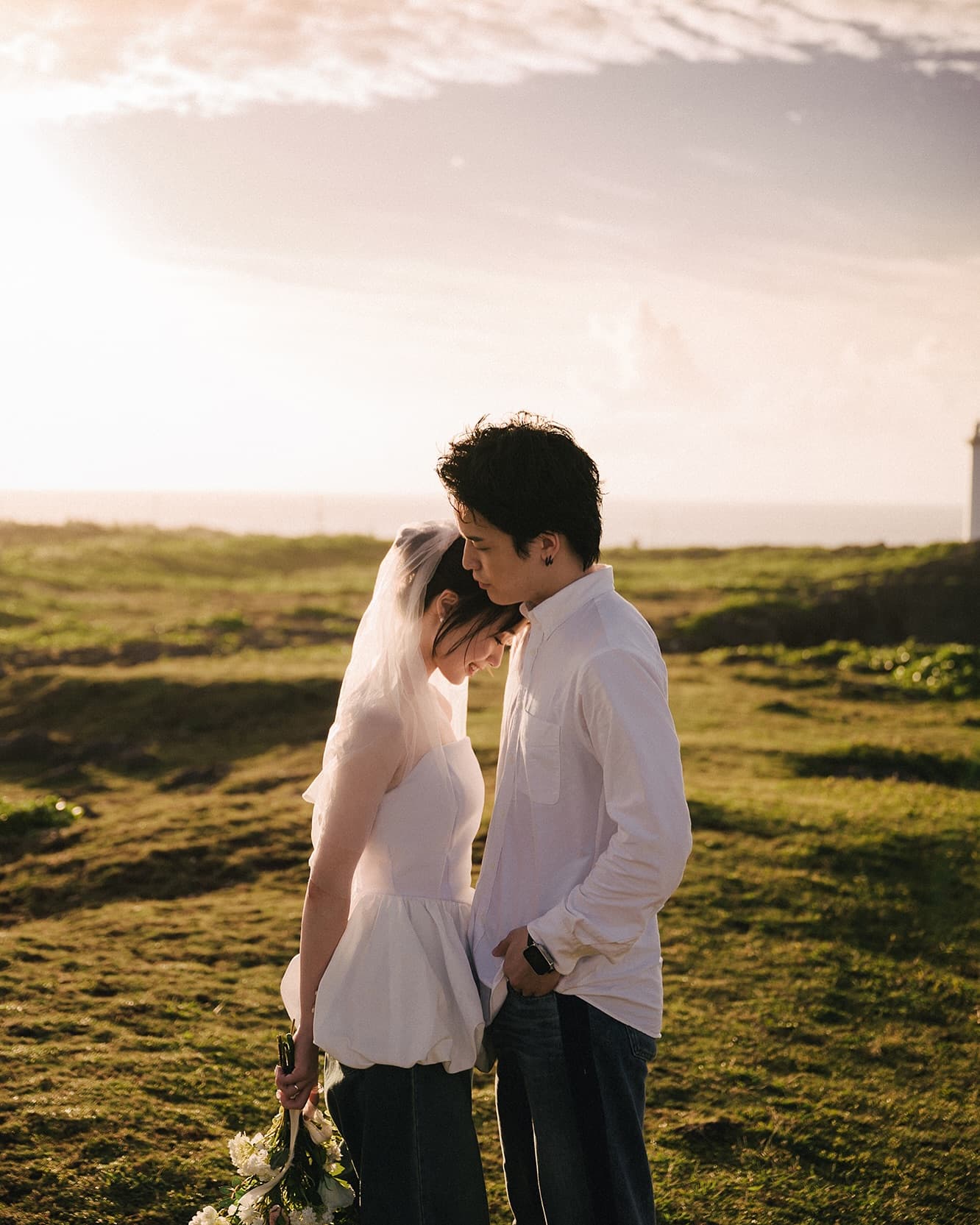 Couple standing close on a backlit grassy field in Okinawa, white tops and denim