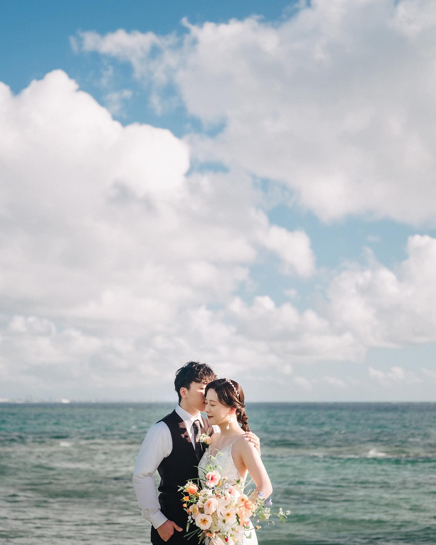 Groom kissing his bride by the sea under a bright blue Okinawa sky