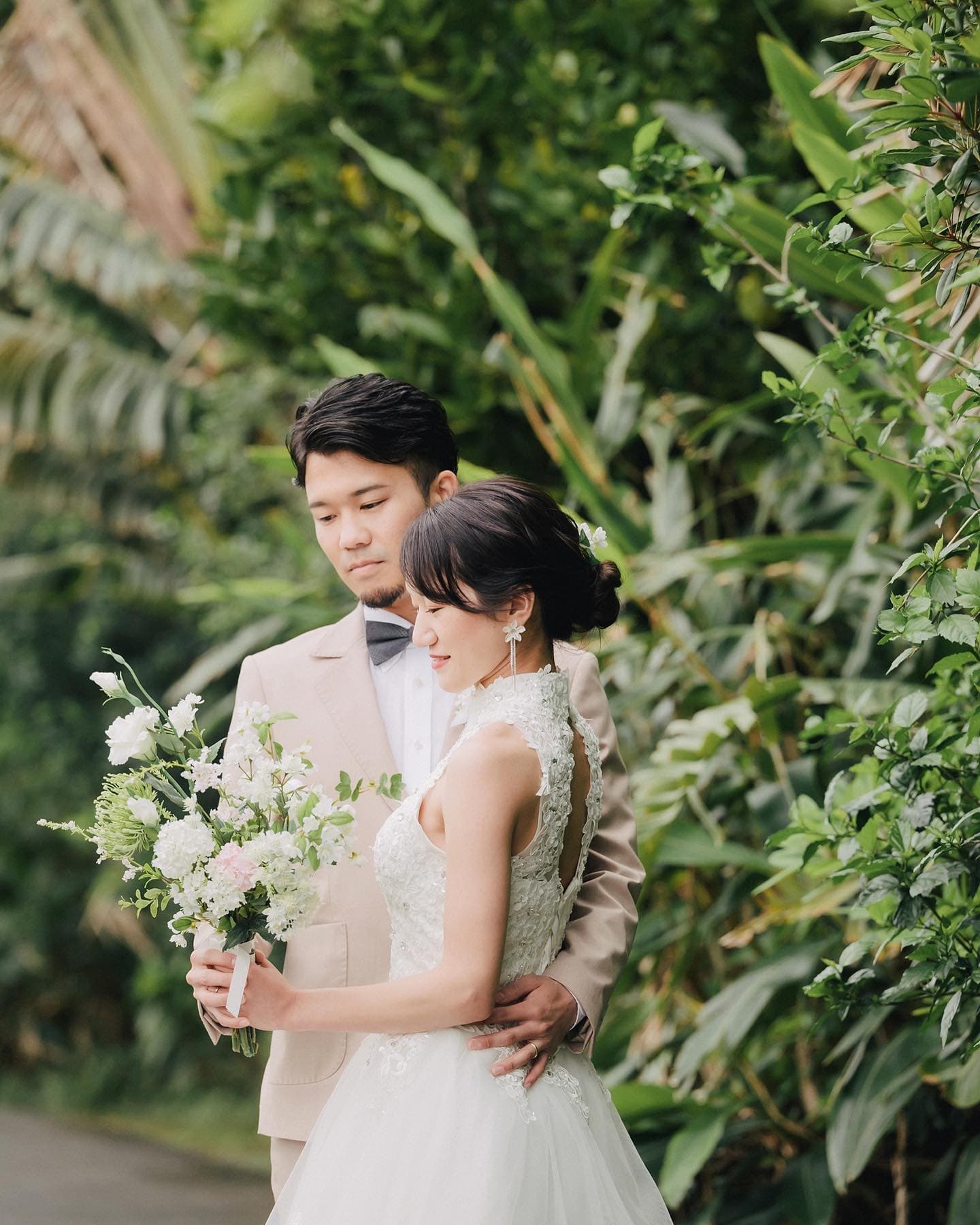 Surrounded by tropical greenery in Okinawa, a groom stands close behind his bride