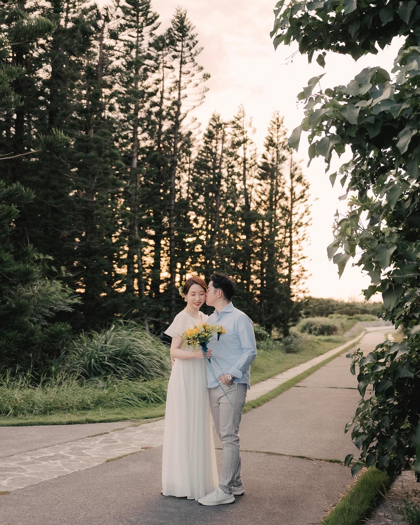 Groom kissing the bride temple on a pine-lined road at dusk