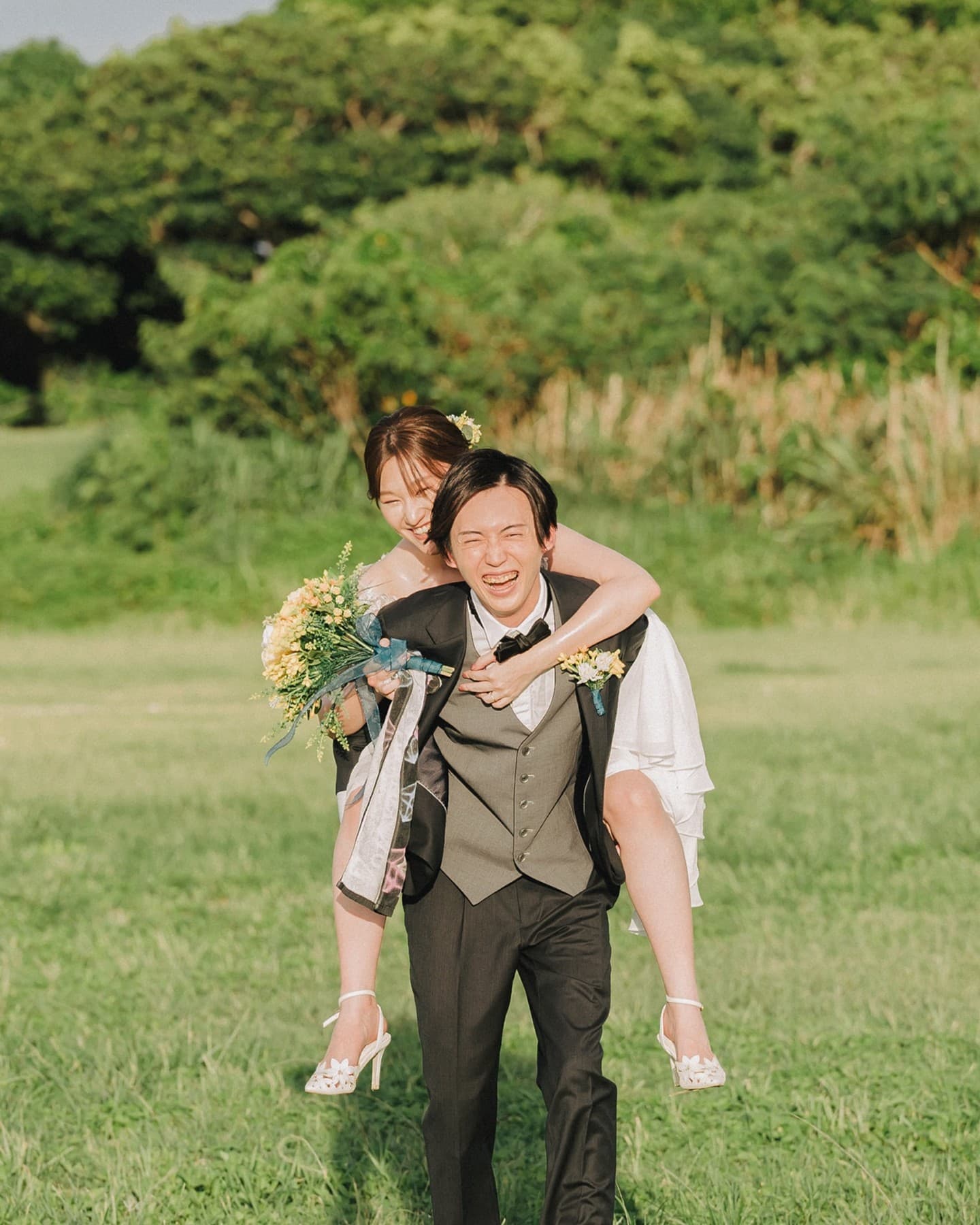 Groom laughing as he piggybacks the bride across a green field in Okinawa.