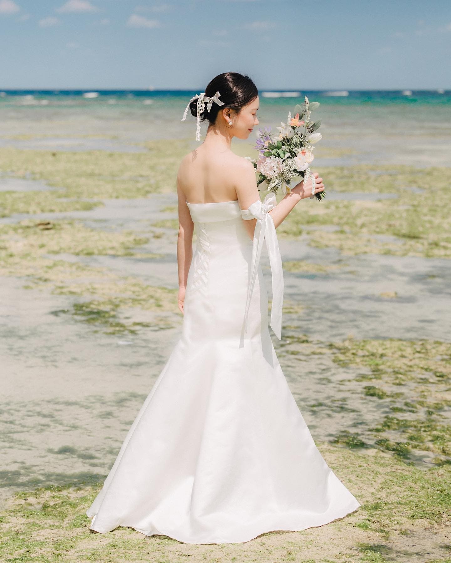 Bride from behind in a mermaid dress holding a bouquet by a shallow Okinawa reef