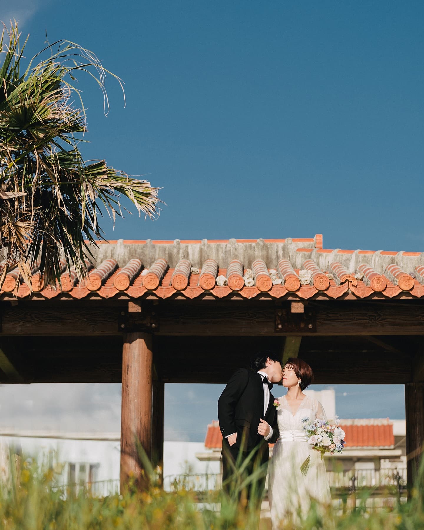 Groom kissing his bride's cheek at a red-tiled pavilion in Okinawa
