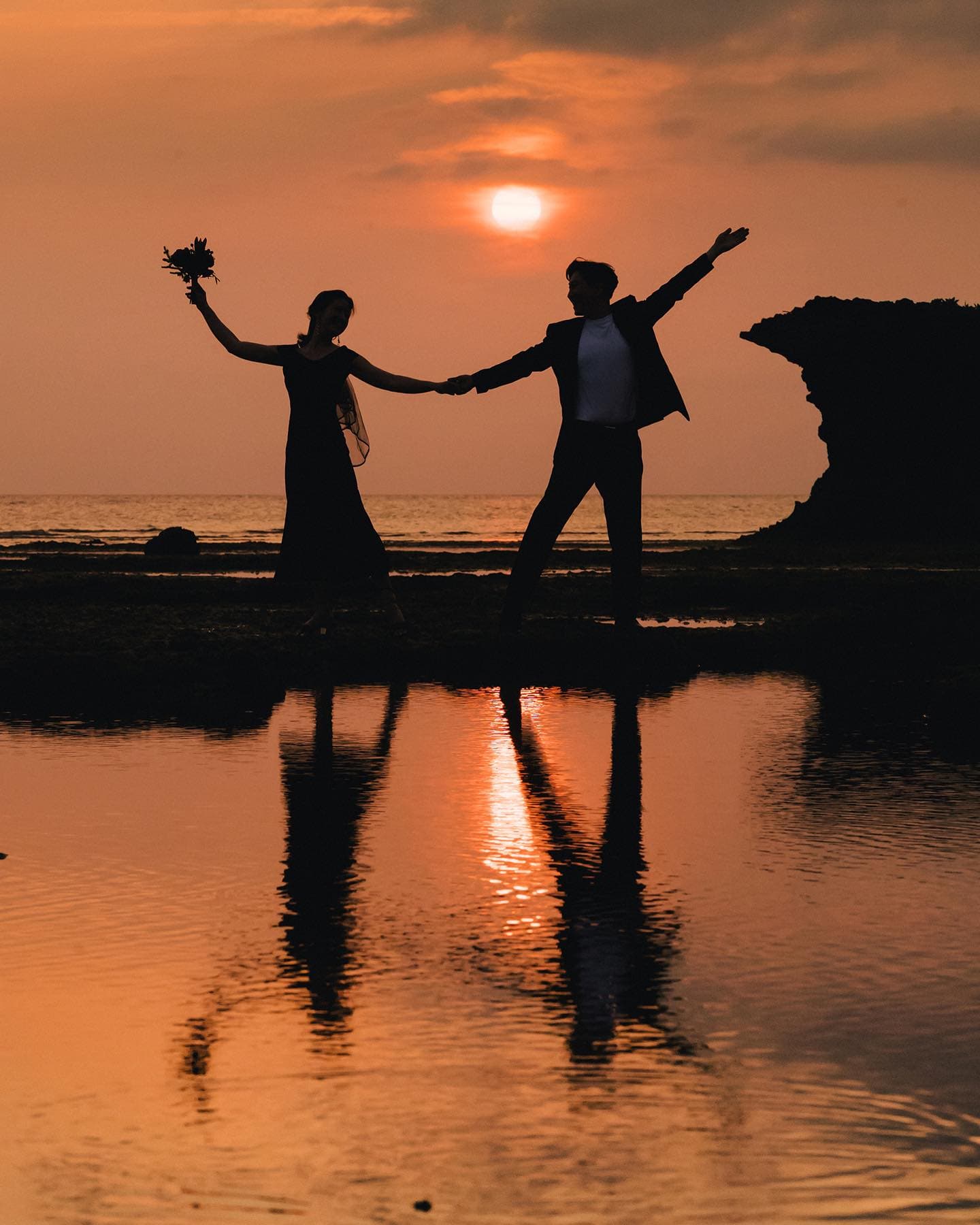 Silhouetted couple with arms spread before an orange sunset, Okinawa