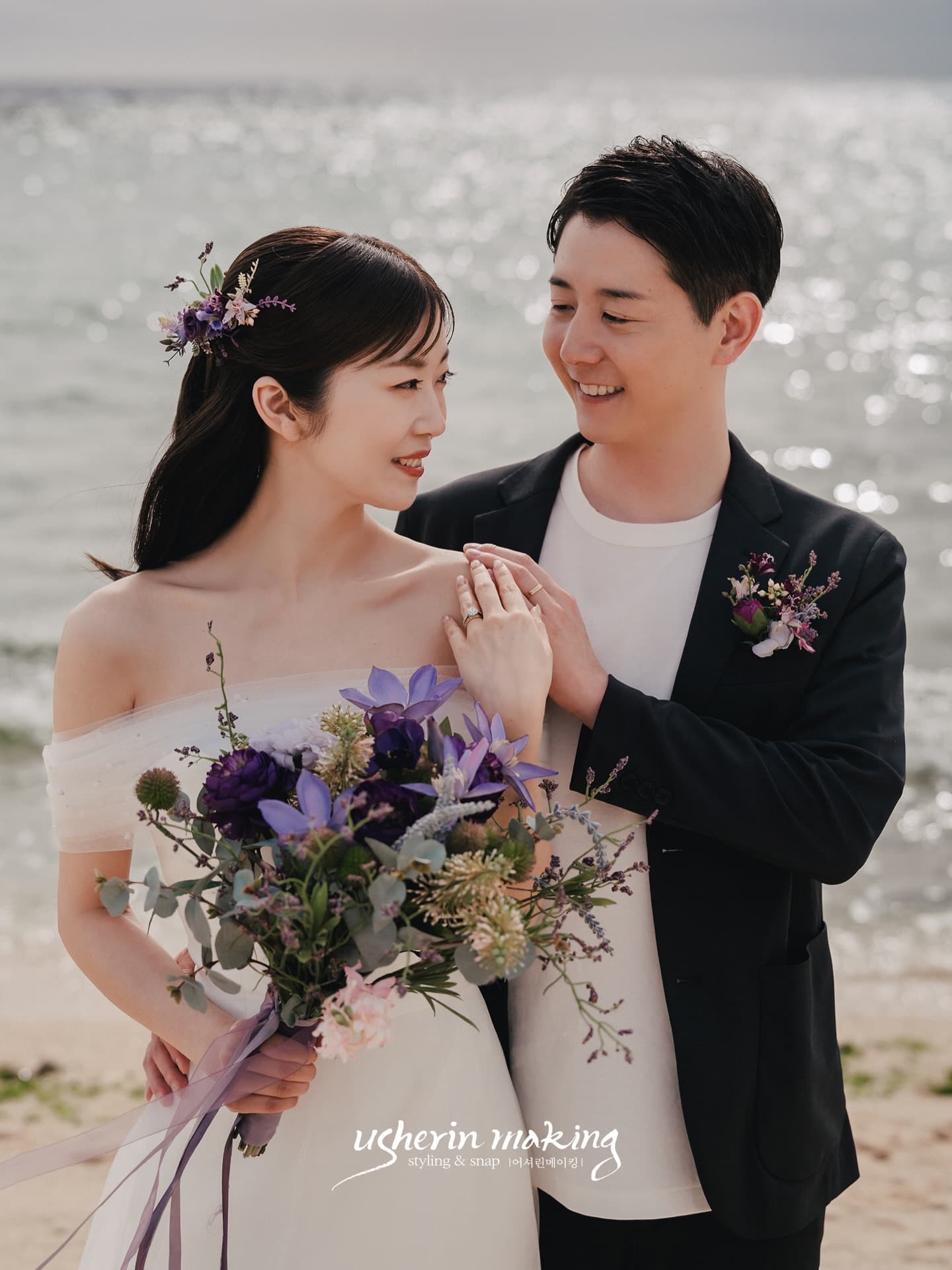 きらめく海を背に見つめ合って微笑む新郎新婦と紫の花束、沖縄ビーチ