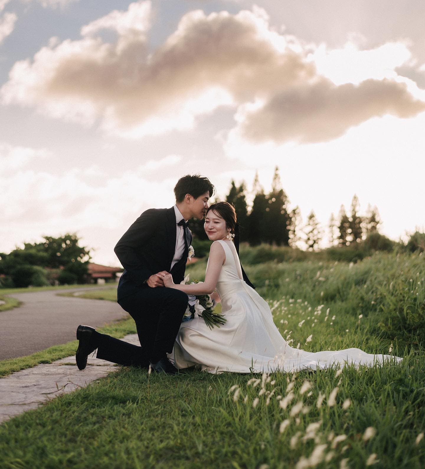 Groom kneeling to kiss his bride cheek in a golden Okinawa meadow