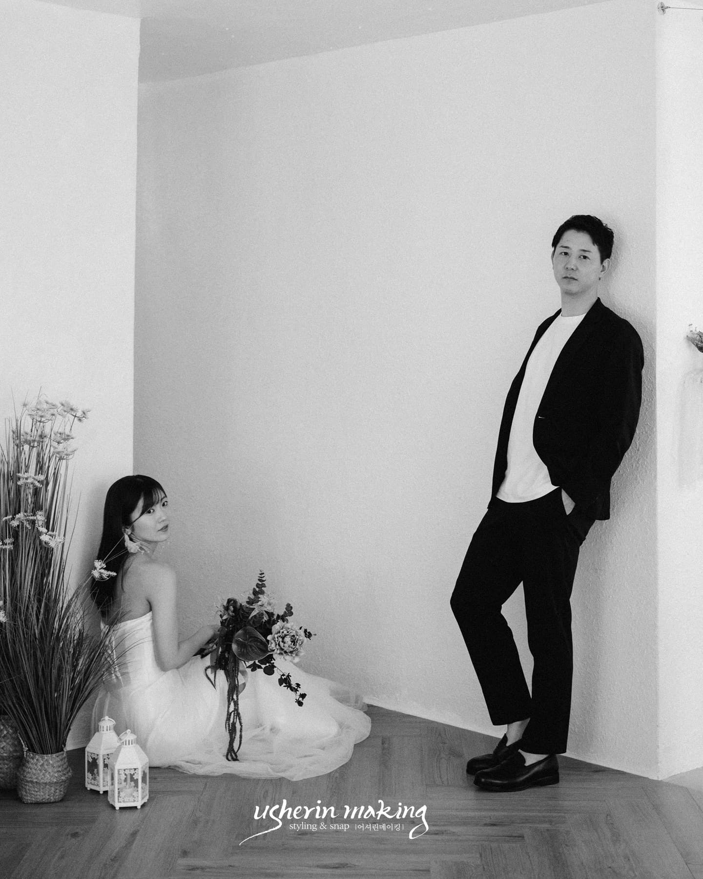 Monochrome studio shot of the bride seated on the floor and the groom leaning on a white wall