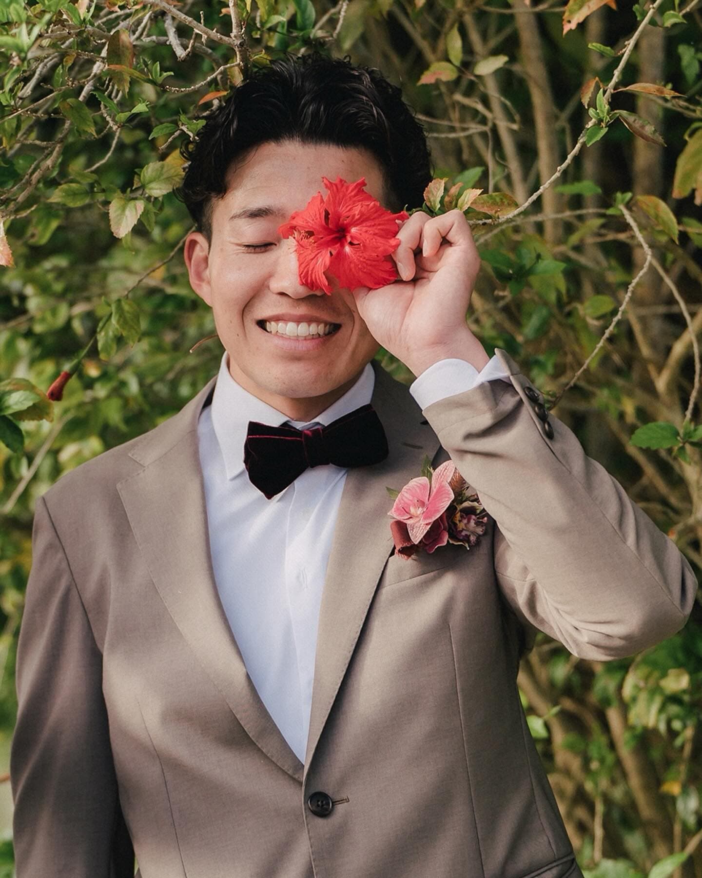 Groom in a beige suit smiles with a hibiscus over one eye in Okinawa