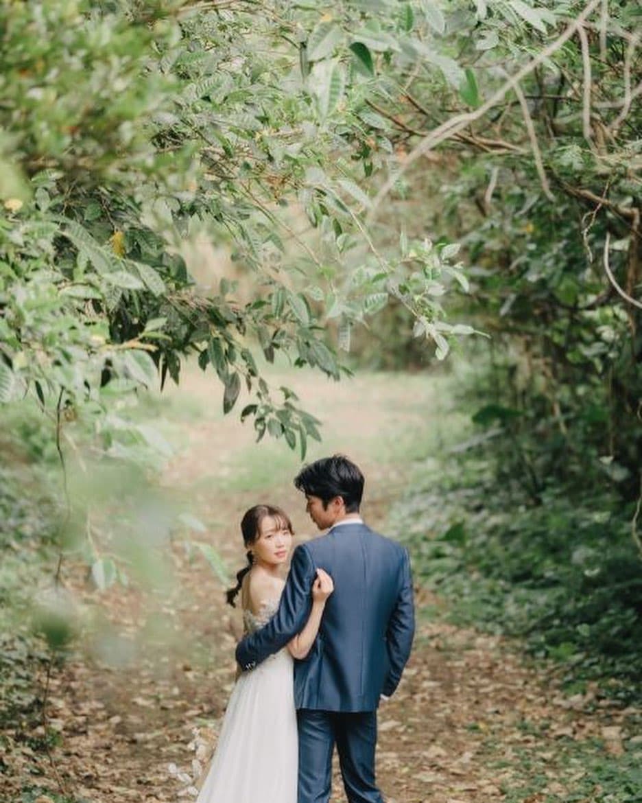 Bride leaning on the groom's arm along a green forest path in Okinawa