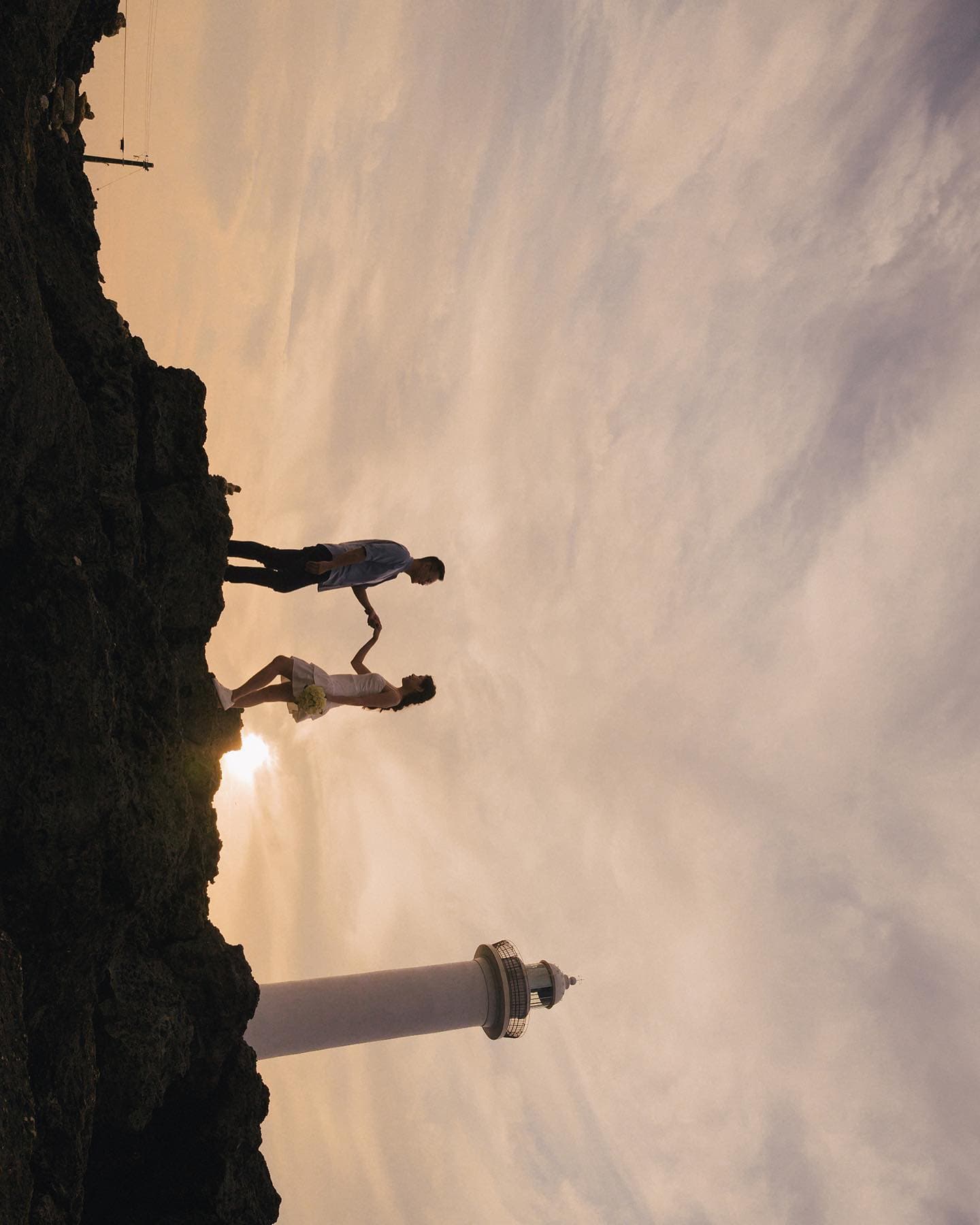 A couple crossing rocky ground near a white lighthouse under a sunset sky in Okinawa