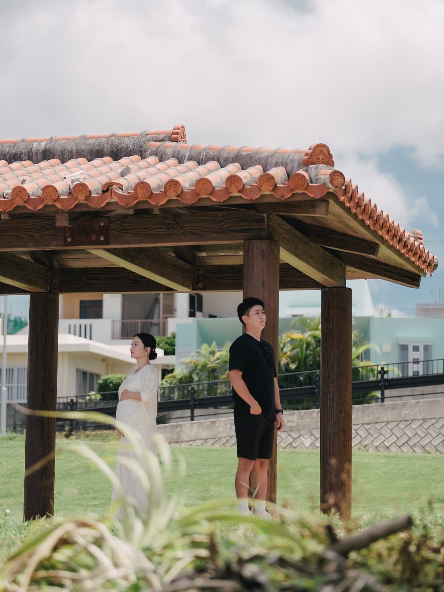 赤瓦の東屋の下にたたずむ白いワンピースの妊婦と夫、曇りの沖縄