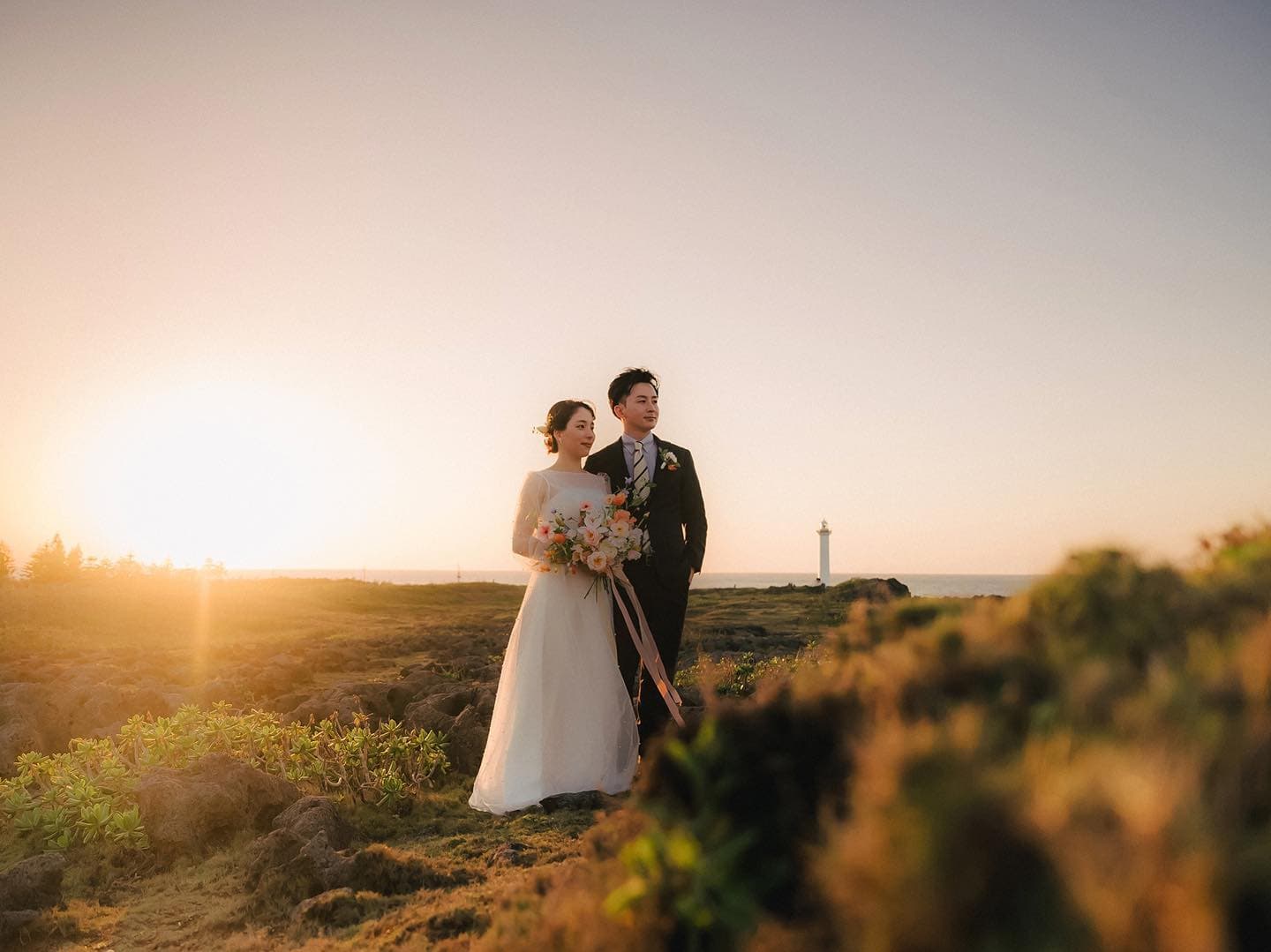 Bride and groom on a sunset hill with a distant lighthouse in Okinawa