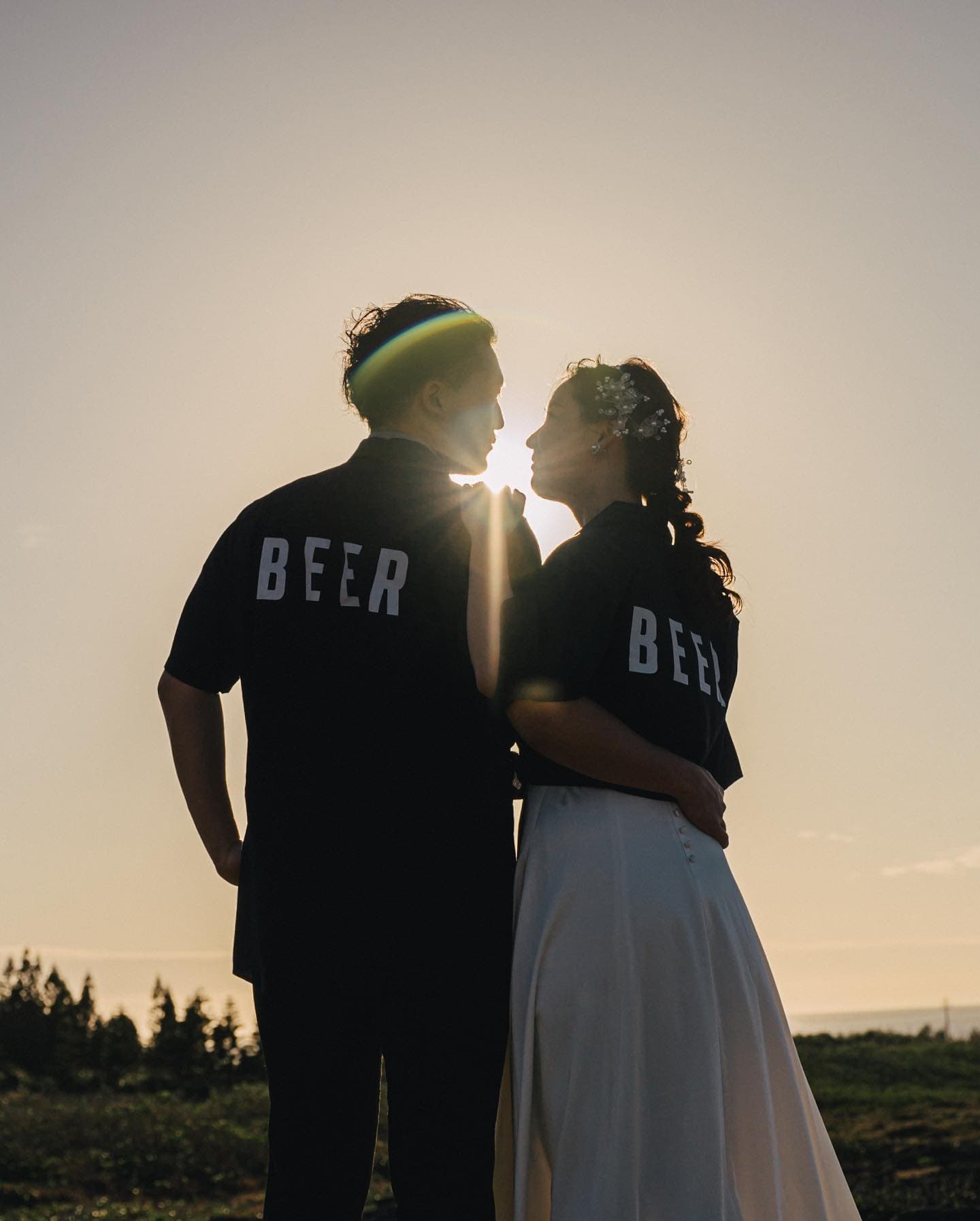 지는 해를 사이에 두고 마주 선 BEER 셔츠 커플의 실루엣