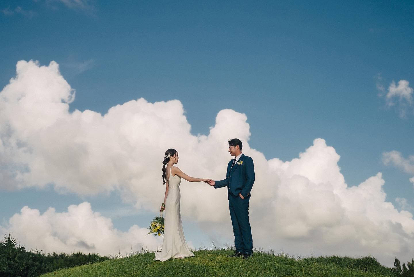 Bride and groom join hands on a hill under towering clouds in Okinawa