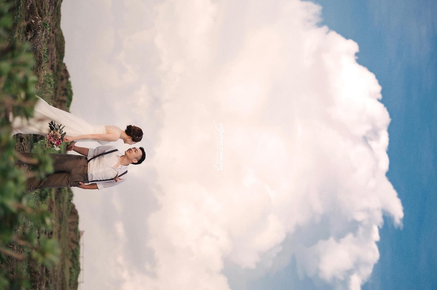 青空と大きな雲を見上げる花嫁と新郎、緑に囲まれた沖縄の丘