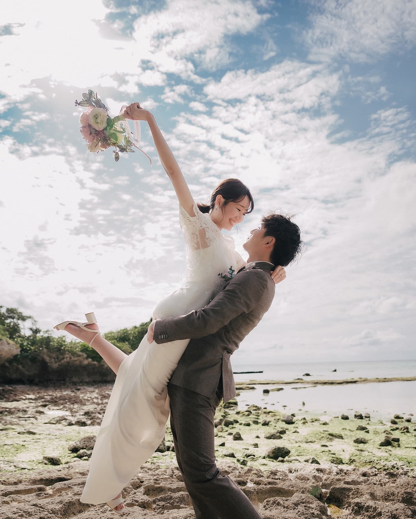 青空の下、花嫁を抱き上げる新郎とブーケを掲げる花嫁、沖縄のビーチ