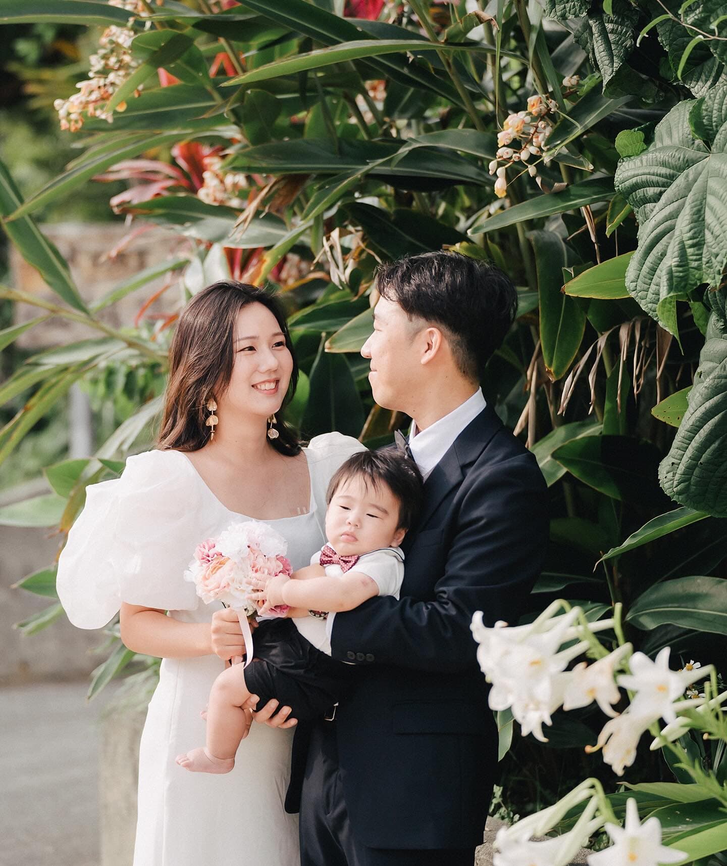Parents in white dress and suit cradling their baby amid tropical blooms in Okinawa