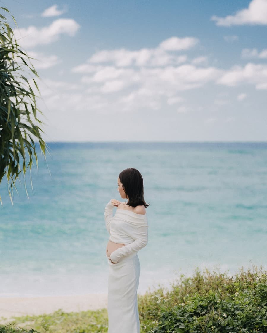 Pregnant woman in white cradling her belly before Okinawa's blue sea and sky