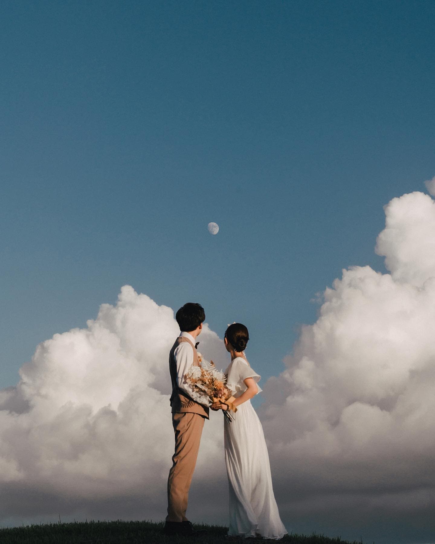 Couple facing each other on a hilltop under a small moon and towering clouds, Okinawa