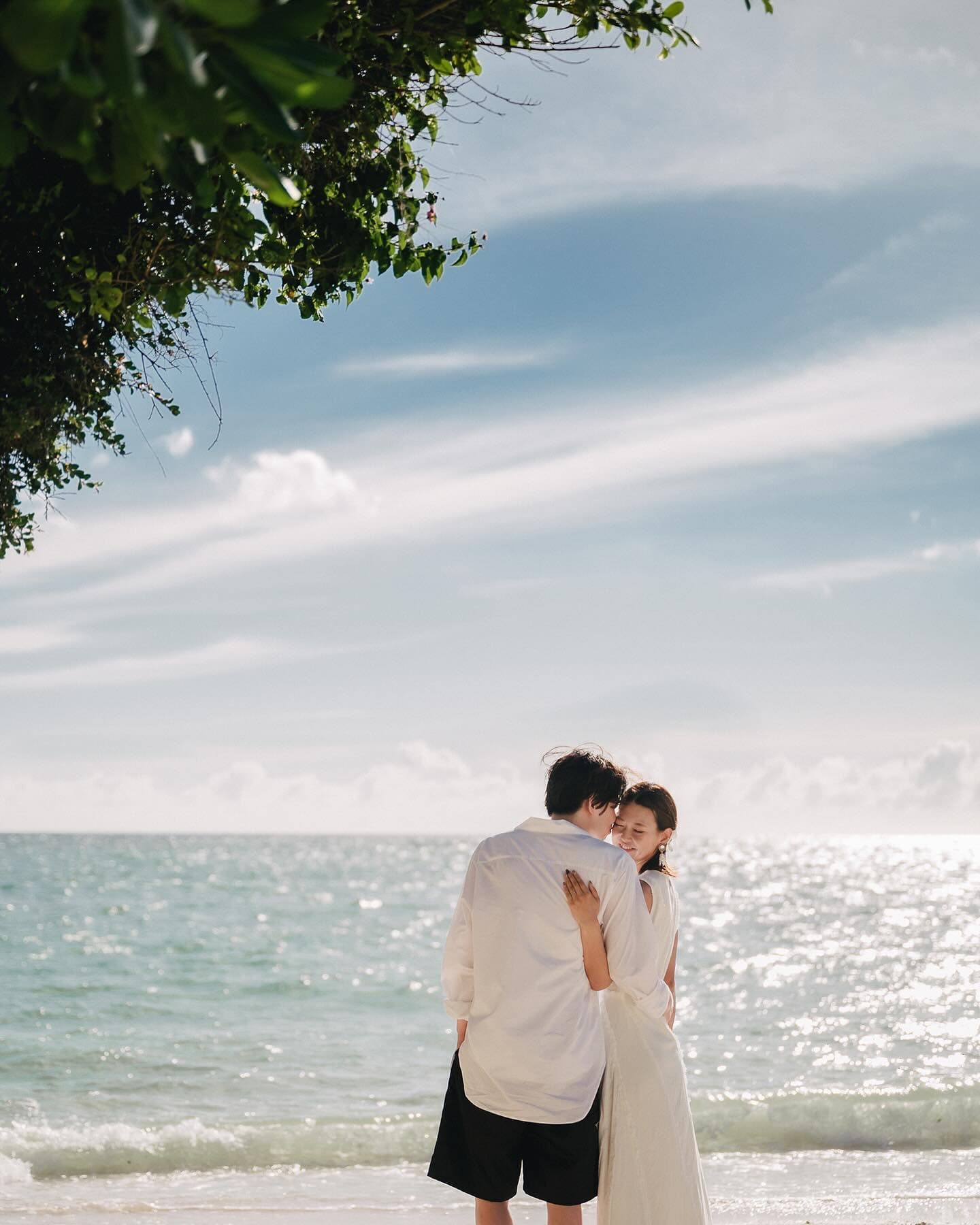 Couple embracing on a sunlit Okinawa beach beneath overhanging leaves