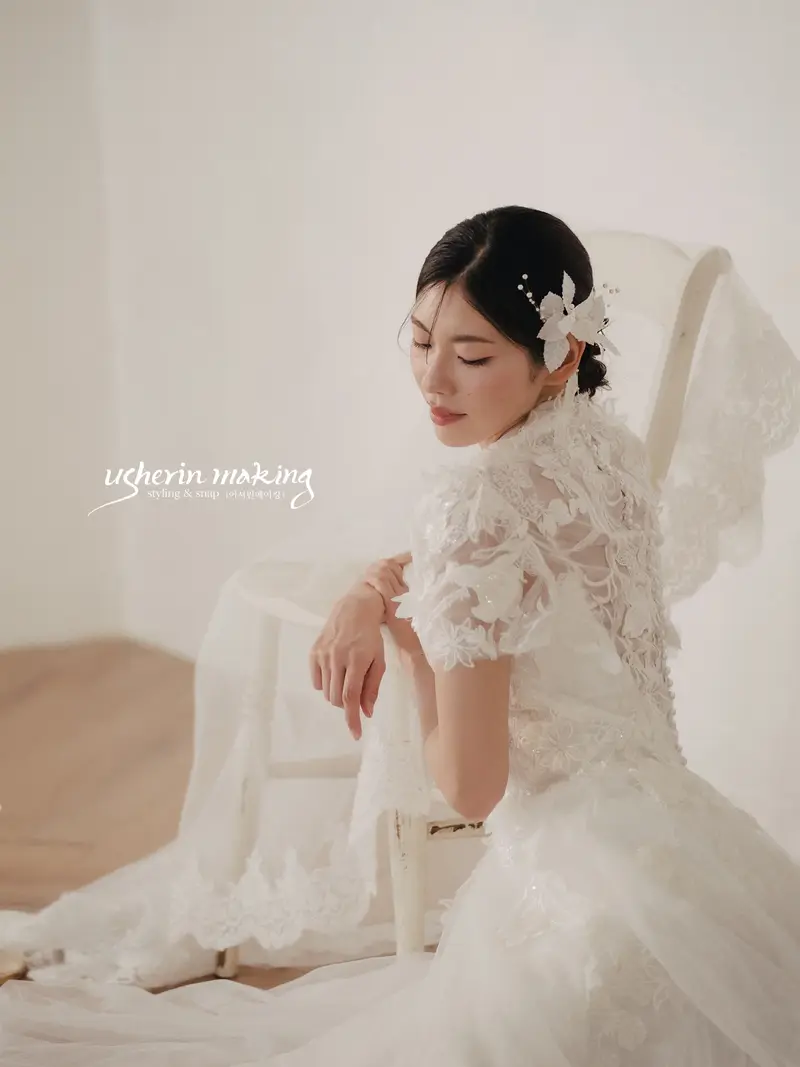 Bride in a lace gown resting on a chair with eyes lowered, in a warmly lit studio