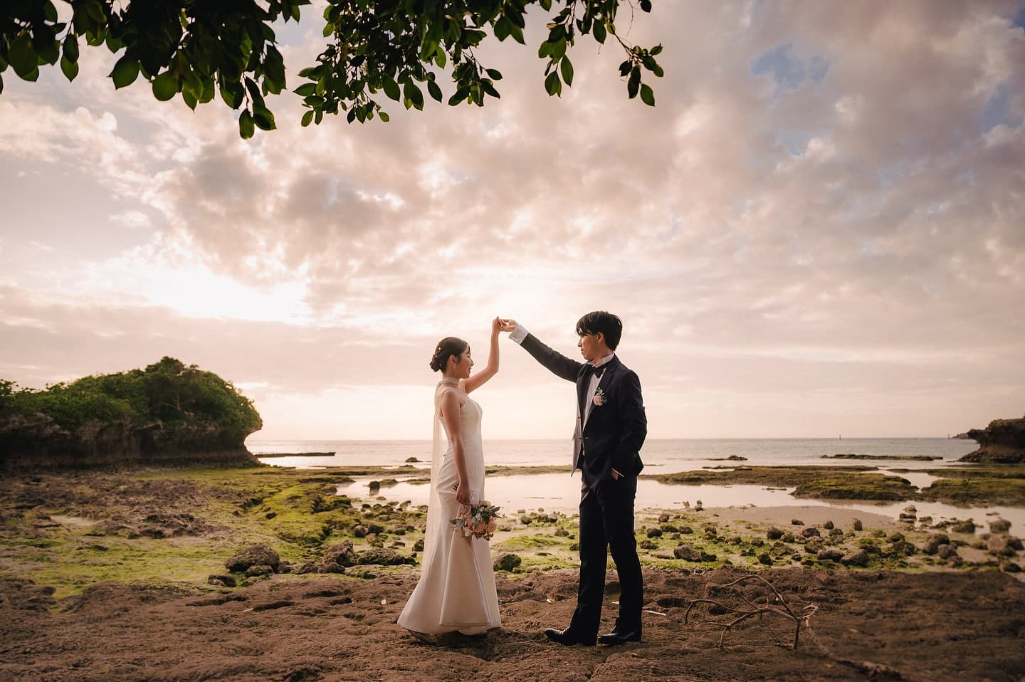Groom raising the bride's hand as if to dance on a rocky shore at sunset in Okinawa